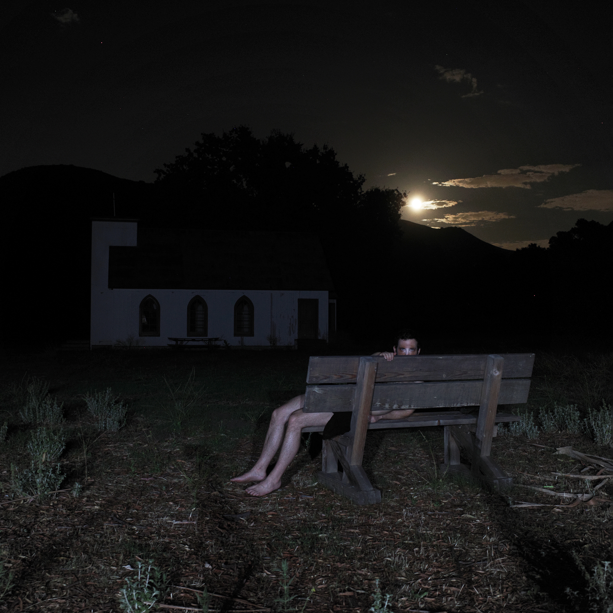 Image of a pale figure reclined on a bench in moonlight with a church behind them peering at the camera.